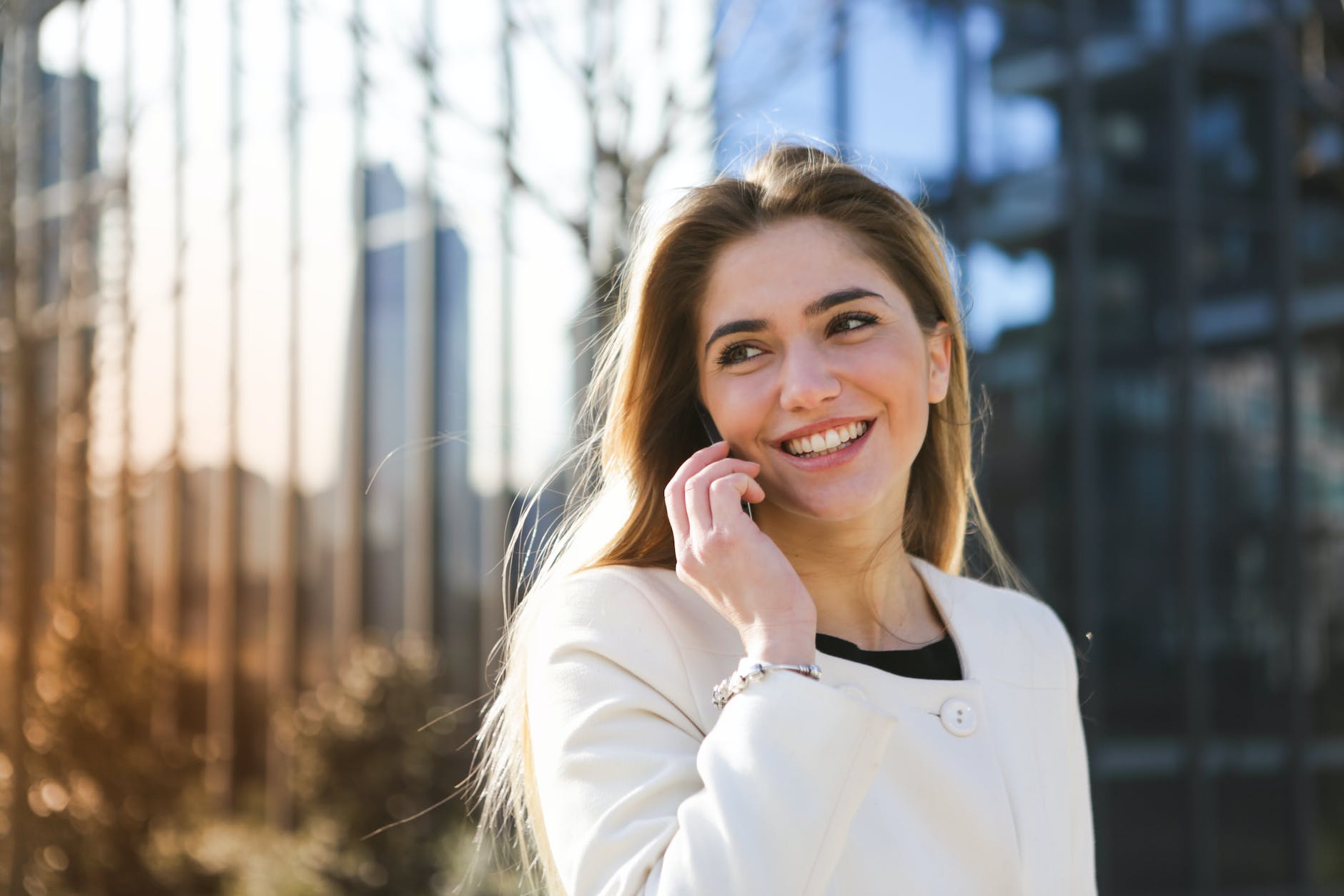 selective focus photo of woman in white long sleeve coat smiling while talking on the phone