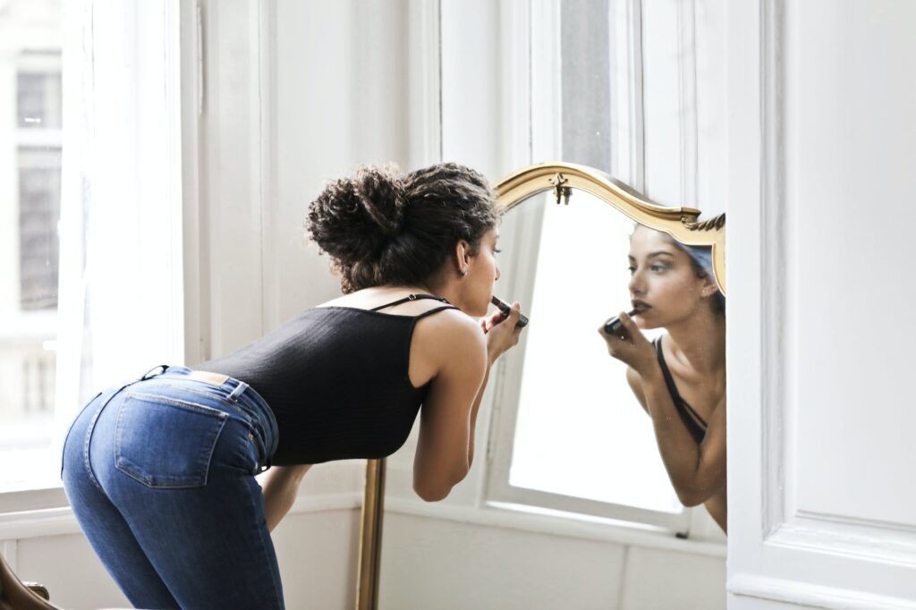 photo of woman in black tank top and blue denim applying lipstick in front of mirror