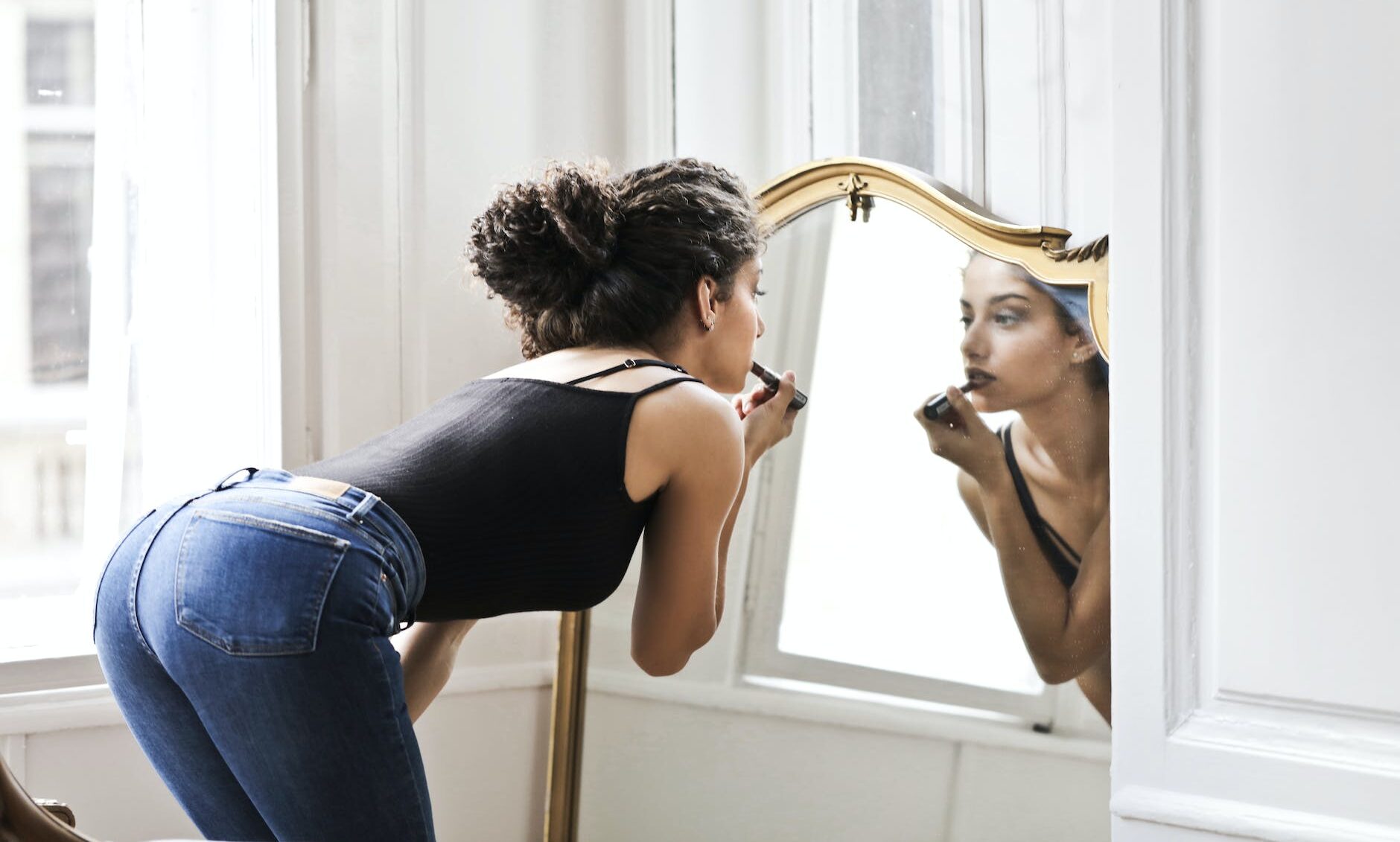 photo of woman in black tank top and blue denim applying lipstick in front of mirror